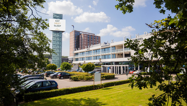 Vooraanzicht van Van der Valk Hotel Eindhoven met groene tuinen en blauwe lucht.